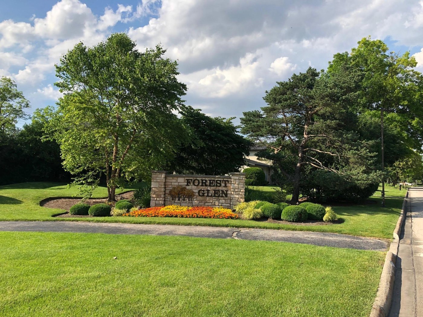 Forest Glen entrance in summer, with green lawns, mature trees, and marigolds blooming at the monument